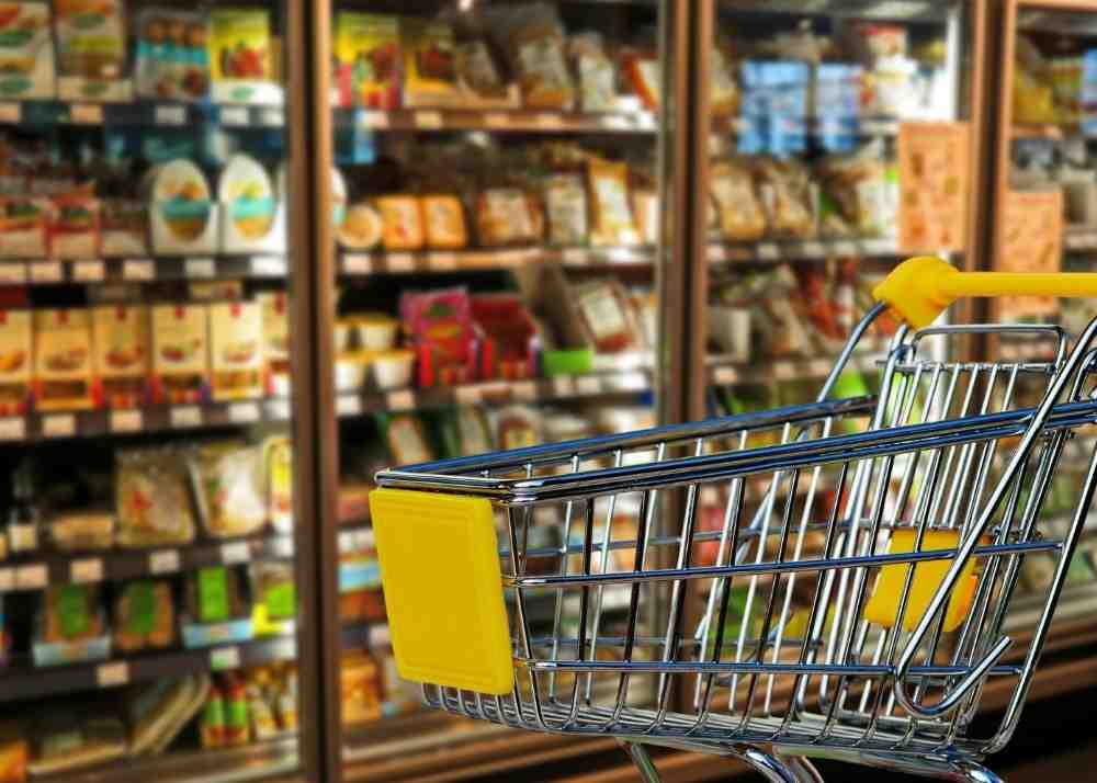 A shopping cart in front of an aisle of food at a grocery store.